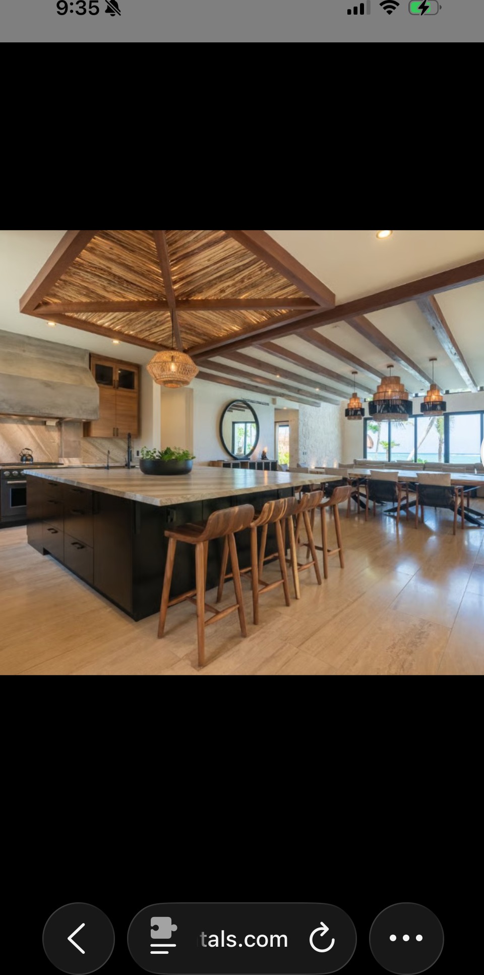 The villa\u2019s great room: a large marble kitchen island with bar stools, exposed beam ceiling with a palapa-style inset, and a long dining table visible beyond, set for a group.