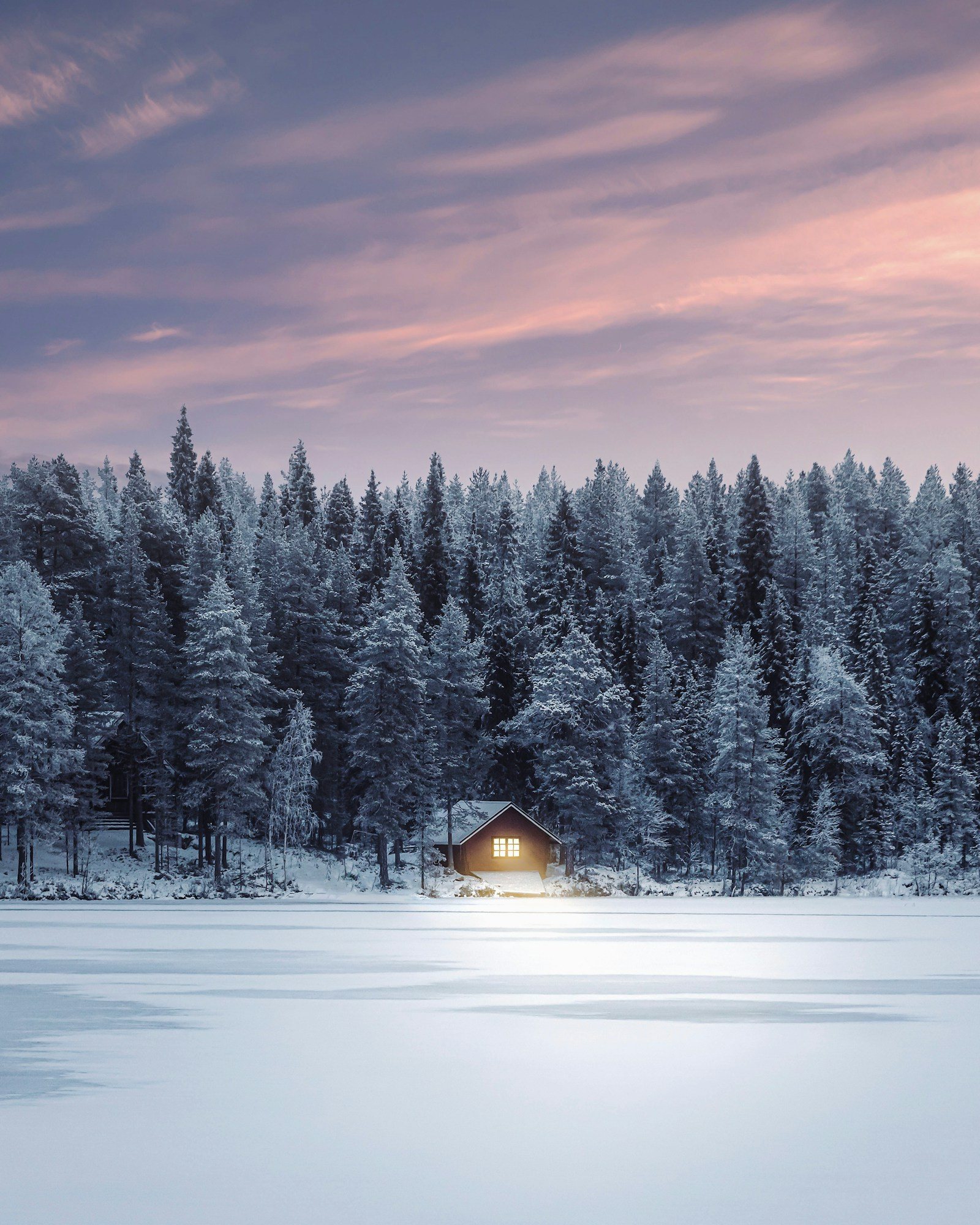 A small log cabin with lit windows beside a frozen lake and snowy pine forest